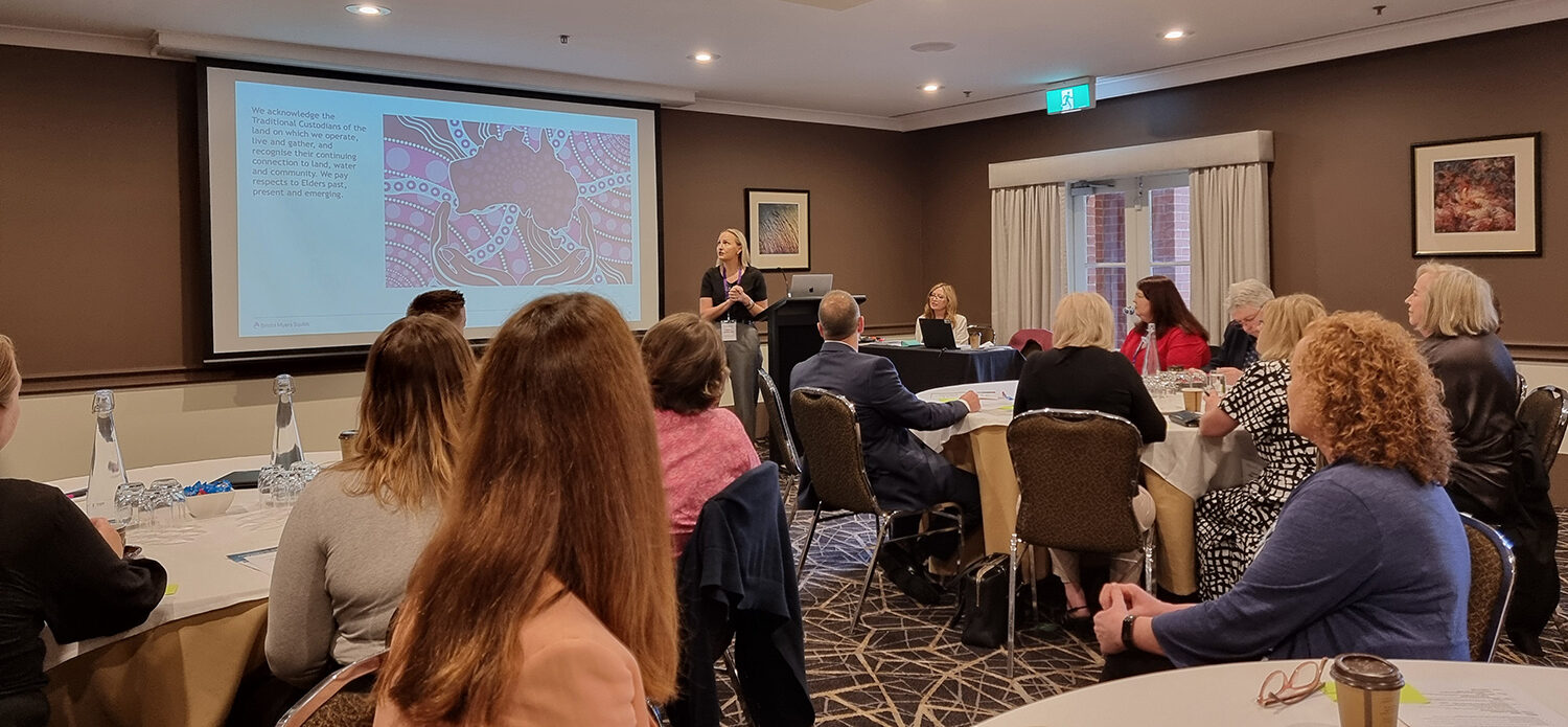 Image is a photo of a group of people sitting around several round white tables. They are looking at a presenter standing between a poduim and a large white screen with an image projected on it. The walls of the room are brown and the ceiling and curtains are white.