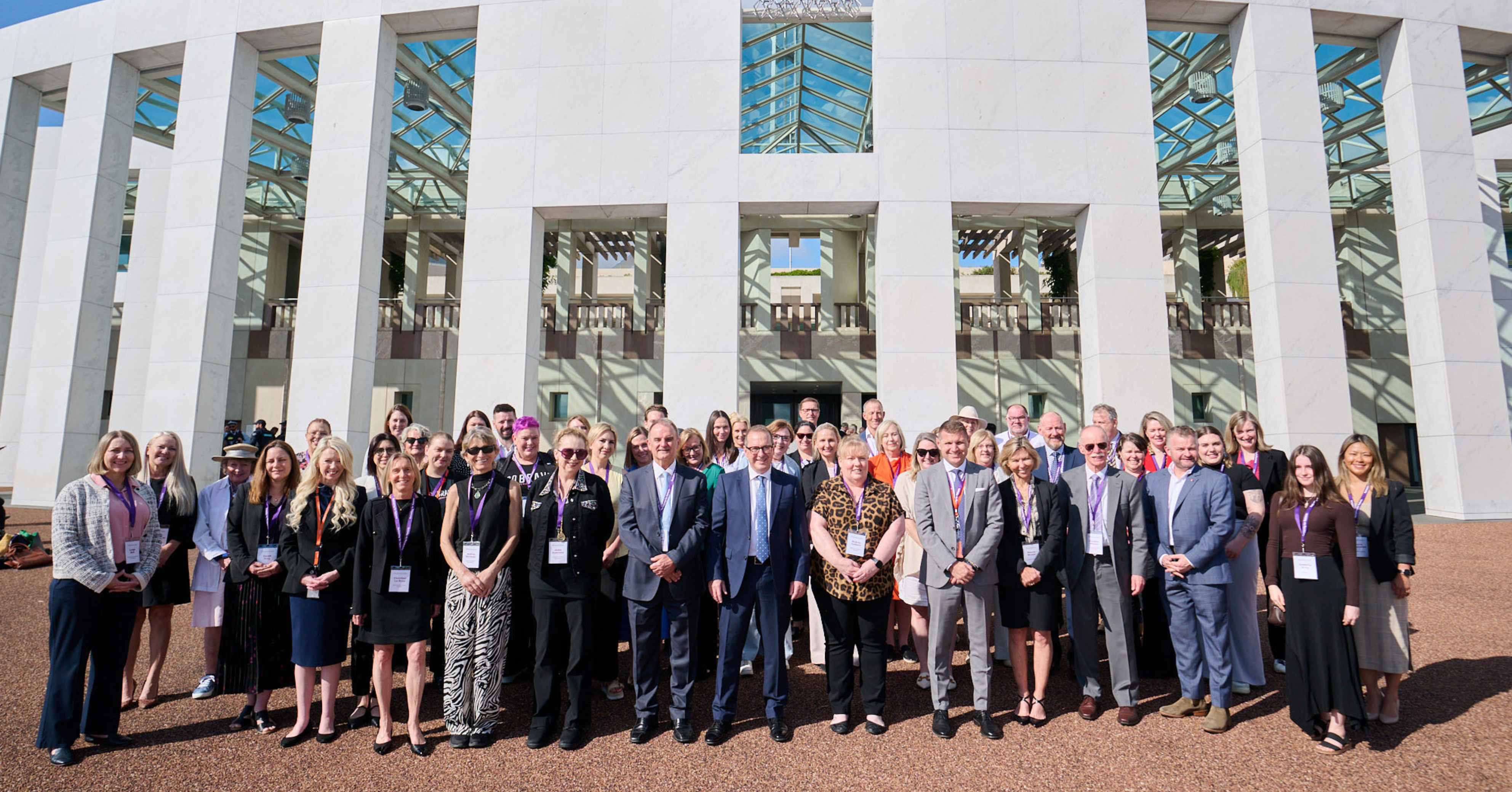 Image is a photo of all the attendees and organisers of the BMS Patient and Parliament Summit 2024 standing outside of Parliament House, Canberra on a sunny day. The building facade is curved and white with tall rectangular pillars. The metal-frame Austrailan Coat of Arms sits on the roofline of the building and the sky is visible through it.