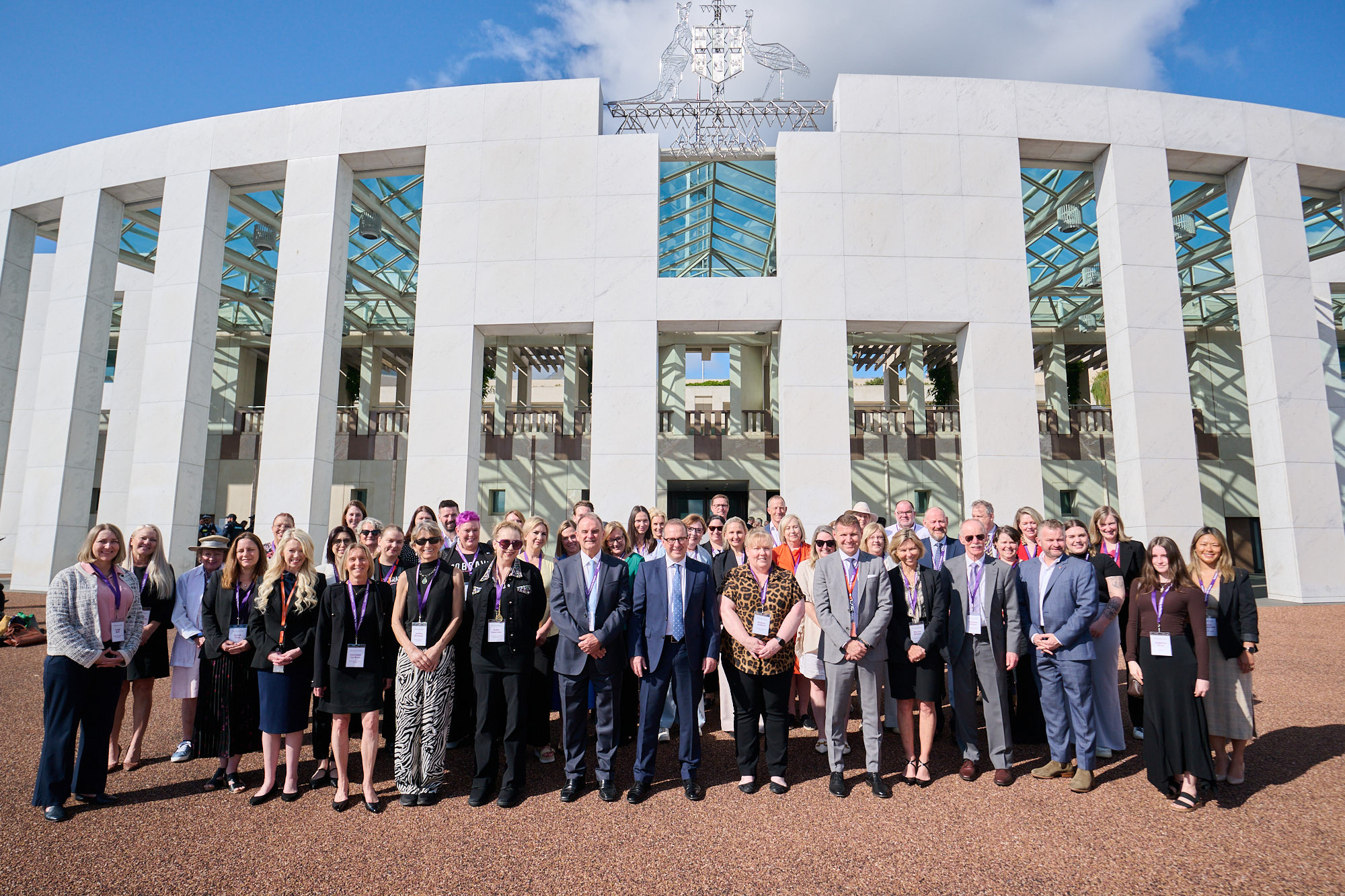 Image is a photo of all the attendees and organisers of the BMS Patient and Parliament Summit 2024 standing outside of Parliament House, Canberra on a sunny day. The building facade is curved and white with tall rectangular pillars. The metal-frame Austrailan Coat of Arms sits on the roofline of the building and the sky is visible through it.